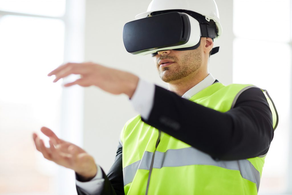 Mann trägt Warnweste und Schutzhelm. Hat VR-Brille auf und bewegt die Hände.